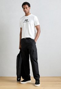 T-shirt blanc à manches courtes avec logo noir, associé à un pantalon cargo noir ample avec des poches latérales. Baskets noires avec semelles blanches.