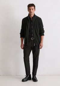 Donkergroene rits-trui over een zwart shirt, gecombineerd met donkere broek en zwarte veterschoenen. De textuur is geribbeld; het ontwerp is modern en slim fit.