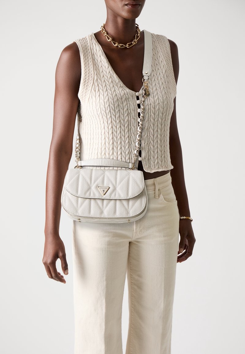 Personne à la peau foncée portant un gilet en tricot sans manches crème, un pantalon crème, un collier chaîne doré, et portant un sac bandoulière blanc matelassé Guess.