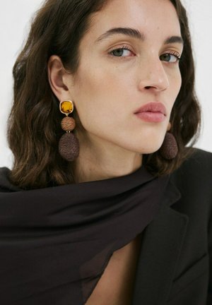 Femme aux cheveux bruns courts portant de grandes boucles d'oreilles pendantes marron et ambre, un blazer noir et une écharpe sombre et transparente.