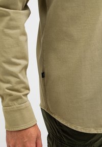 Olivenfarbendes Baumwollhemd mit langen Ärmeln und einer dezenten Textur. Verfügt über eine sichtbare Naht am Saum und ein kleines schwarzes Etikett an der Seite.