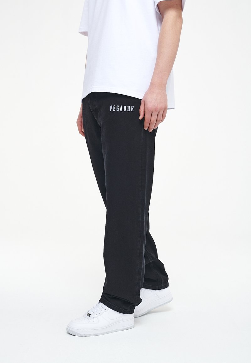 Svarta denimbyxor med "PEGADOR" broderat i vitt på vänstra låret, kombinerat med en vit t-shirt och vita sneakers.