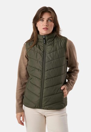 Femme portant un gilet sans manches matelassé vert olive sur un pull marron à manches longues et un pantalon beige clair, debout avec une main dans la poche du gilet.