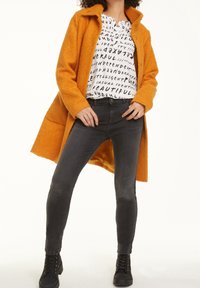 Oranger Teddymantel mit breitem Kragen, schwarz-weißes Grafikhemd mit Text, dunkelgraue Skinny Jeans und schwarze Schnür-Ankle Boots.