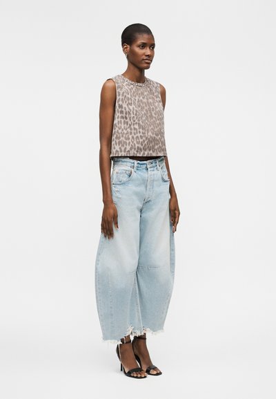 Γυναίκα που φοράει αμάνικο τοπ με λεοπάρ μοτίβο, φαρδιά ανοιχτό μπλε τζιν με ξεφτισμένες άκρες και μαύρα ανοιχτά πέδιλα με ψηλό τακούνι, στέκεται πλαγίως.