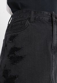 Černá džínsová sukně s poškozenými místy, vypasovaným pasem a standardními kapsami. Tkanina má texturovaný povrch, který zvyšuje její drsný vzhled.