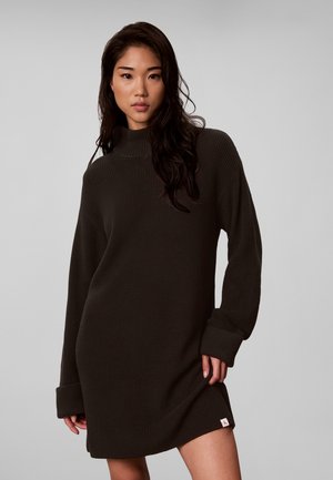 Donna che indossa un abito maglione scuro a maniche lunghe con collo alto, in piedi davanti a uno sfondo chiaro e uniforme.