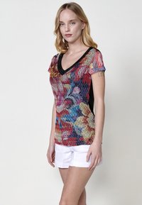 Top de escote en V multicolor que presenta un patrón floral texturizado, mangas cortas y paneles laterales negros. Combinado con pantalones cortos blancos.