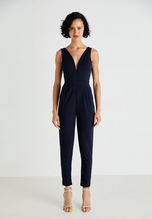 Combinaison bleu marine avec un décolleté en V profond, une taille cintrée et des jambes fuselées. Le matériau semble lisse et extensible ; portée avec des talons à brides.