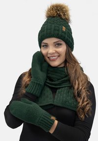 Groene gebreide beanie met een nepbont pompon, bijpassende handschoenen en sjaal, met textuurpatronen en een leren logo-accent.