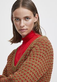Pull en crochet marron et rouge avec un design en V, présentant un motif texturé, superposé sur un haut à col roulé rouge.