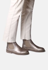 Bottines Chelsea en bronze métallique avec des panneaux élastiques sur les côtés, texture lisse et bout rond, associées à un pantalon beige cropped.