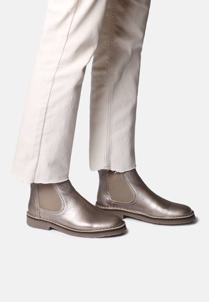 Bottines Chelsea en bronze métallique avec des panneaux élastiques sur les côtés, texture lisse et bout rond, associées à un pantalon beige cropped.