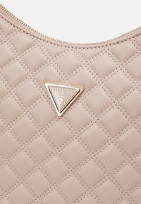 Sac à main matelassé beige clair avec des coutures diagonales et une plaque triangulaire dorée avec le logo Guess près de la fermeture éclair supérieure.