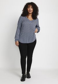 Langærmet v-hals bluse i blå med et struktureret diamantmønster, parret med sorte slim-fit bukser og sorte sko.