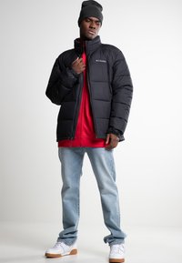 Sort pufferjakke med quiltet struktur, høj krave og lynlås. Båret over en rød hoodie, kombineret med lyseblå jeans og hvide sneakers.