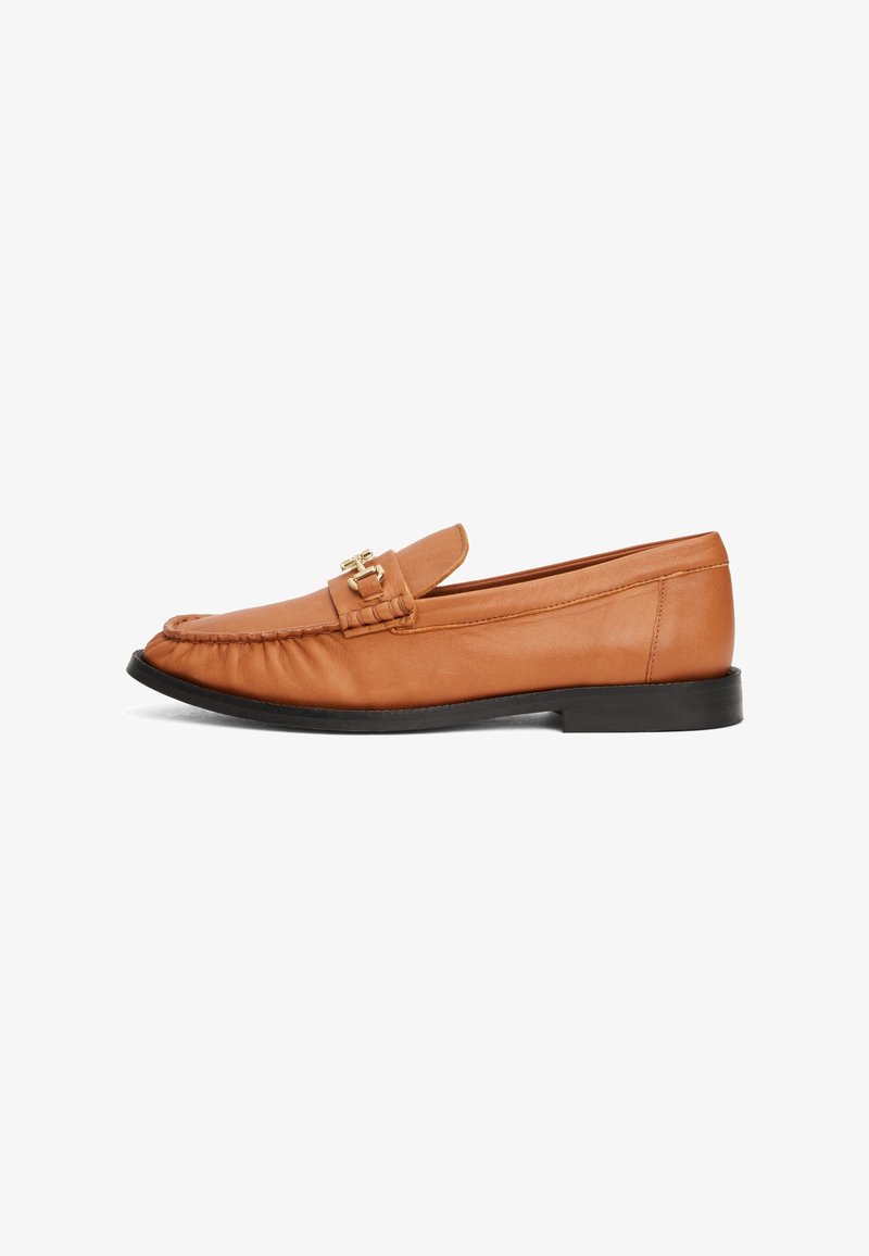 Mocassin en cuir marron avec un bout cousu, semelle plate noire et accent en métal doré à l'avant. Texture lisse, forme classique.