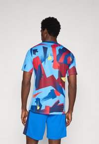 Rövid ujjú, kék sportpóló, merész, piros, sárga és sötétkék absztrakt mintázattal. Lélegző, könnyű anyagból készült.