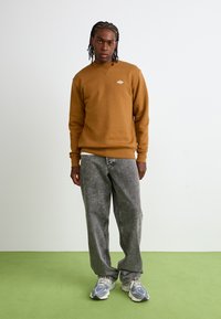 Felpa marrone con piccolo logo, pantaloni in denim grigi e scarpe da ginnastica. Il tessuto appare morbido con una vestibilità rilassata. Palette di colori semplice e tenue.