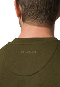 Oliwkowy sweatshirt z okrągłym dekoltem i subtelną teksturą. Posiada haftowaną metkę "PRÓCHNIK" z tyłu blisko szyi.
