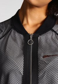 Närbild av en kvinnas nacke och överkropp som bär en svart mesh-bomberjacka med dragkedja och kontrasterande svart krage.