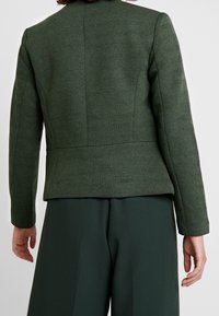 Dos d'une femme portant un blazer cintré en chevrons vert foncé à manches longues et un pantalon large vert foncé sur un fond uni.
