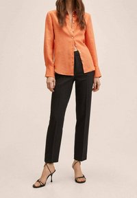 Femme portant un chemisier orange à boutons, un pantalon noir sur mesure et des sandales à talons hauts noires à lanières, debout devant un fond uni.