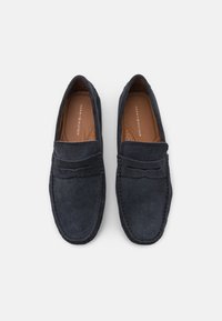 Para czarnych zamszowych mokasynów Tommy Hilfiger z brązowymi, wyściełanymi wkładkami, klasycznym detalem w postaci paska penny, widziana z góry na białym tle.