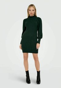 Donna che indossa un vestito a coste verde scuro con maniche a sbuffo lunghe e stivali neri alla caviglia, in piedi su uno sfondo bianco.