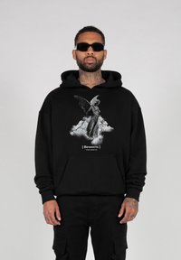 Schwarzer Kapuzenpullover mit Grafikdruck eines Engels auf Wolken, darunter in Weiß textlich hervorgehoben. Das Material scheint weich zu sein, mit einem entspannten Schnitt und einer Fronttasche.