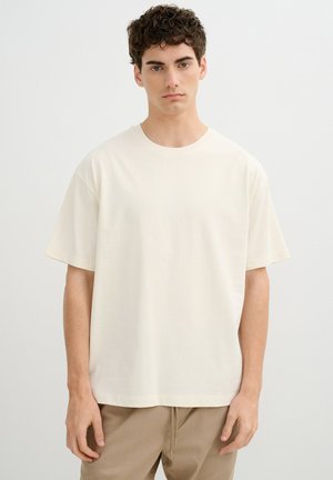 Joven con cabello rizado oscuro, vistiendo una camiseta crema holgada y pantalones beige, de pie contra un fondo claro y liso.