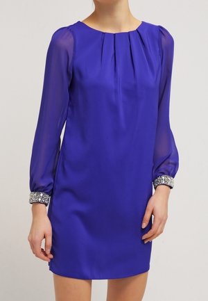 Mujer con un vestido azul real de manga larga translúcida y puños plateados adornados, posando contra un fondo liso.