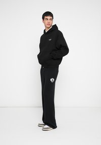 Juodas gobtuvu megztinis su priekiu kišene ir baltu logotipu, derinamas su juodomis ribuotomis sportinėmis kelnėmis, ant kojos esančiu balto kaukolės grafiku.