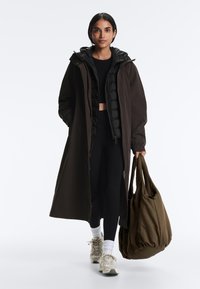 Nő, hosszú sötét kabátot visel fekete ruhán, fehér zoknival, sportcipővel, és jobb kezében egy nagy sötét olívazöld táskát hord.