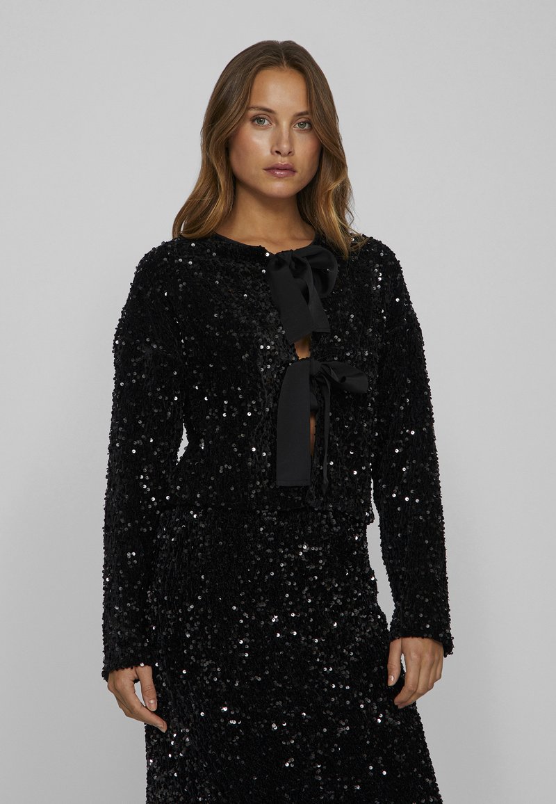 Sorte pailletcroppede jakke med lange ærmer og sløjfeåbning ved halsen. Glat tekstur og shiny finish reflekterer lyset.