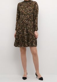 Robe imprimée florale en vert olive avec des manches longues, un col froncé et une jupe à volants. Une paire de talons aiguilles noirs.