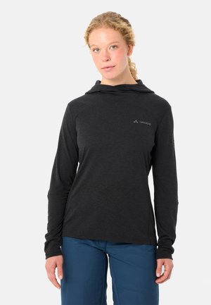 Jonge vrouw met krullend blond haar, gekleed in een zwarte hoodie met lange mouwen en een blauwe broek, kijkt ontspannen naar voren.