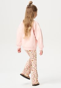 Rosa Sweatshirt mit fallenden Schultern, kombiniert mit rosa gemusterten Schlaghosen mit Blumen- und Fruchtentwürfen; braune Slipper sichtbar.