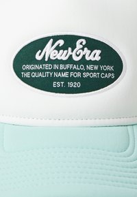Bijela kapa s mint zelenom unutrašnjošću. Ima vezeni ovalni logo s natpisom "New Era" i tekstom ispod. Glatka tkanina s ušivenim detaljima.