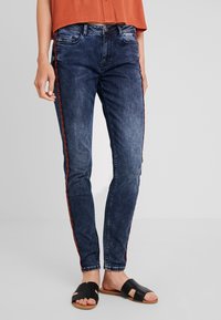 Jeans ajustados de color azul marino con una franja lateral de patrón rojo, diseño de cinco bolsillos y cintura de altura media. Combinados con sandalias negras tipo slip-on.