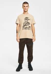 Beżowa grafitowa koszulka z abstrakcyjnym czarnym wzorem i logo, zestawiona z brązowymi spodniami dresowymi z boczną kieszenią na zamek i czarnymi trampkami.