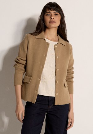 Kobieta w beżowej teksturowanej kurtce zapinanej na guziki, na białej bluzce z kwiatowym haftem i ciemnych spodniach, stojąca na jednolitym tle.