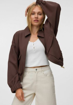 Femme portant une veste courte marron par-dessus un débardeur blanc boutonné et un pantalon beige taille haute, posant avec un bras levé au-dessus de la tête.