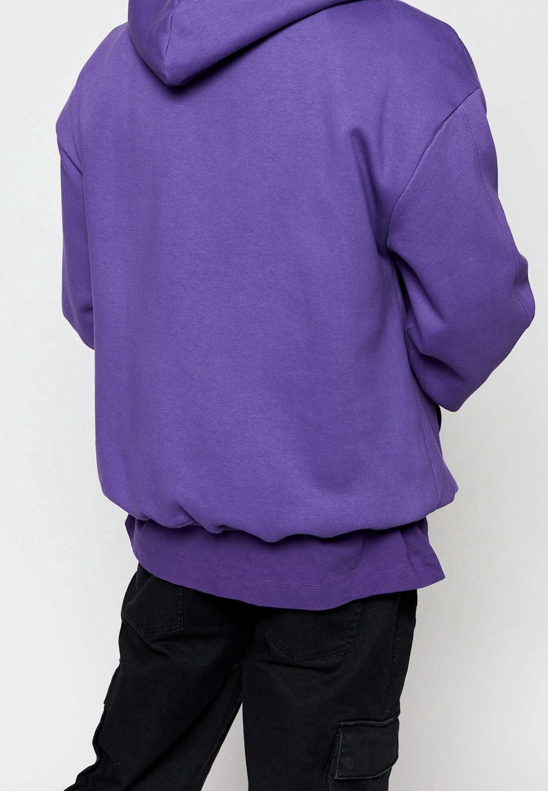 Personne portant un sweat à capuche violet ample et un pantalon cargo noir, vue de dos sur un fond clair uni.