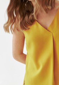 Yellow sleeveless top with a V-neck, featuring soft, smooth fabric and a center pleat, with side slits for added detail.