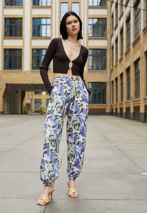 Blusa castanha de mangas compridas com detalhe em forma de buraco, combinada com calças largas e estampadas em tons de verde e azul, e sapatos de salto bejes com tiras.