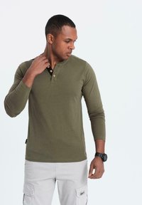 Man i olivgrön långärmad henley-tröja som justerar kragen, bär ljusgrå cargo-byxor och svart armbandsur, ser ner åt höger.