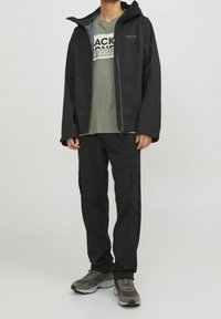 Chaqueta impermeable negra con capucha, forro gris y logo en el pecho, combinada con pantalones cargo negros y zapatillas deportivas grises. Ropa casual para actividades al aire libre.