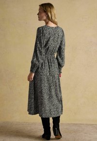 Frau in einem schwarz-weißen, floral gemusterten, langärmligen Midikleid und schwarzen Stiefeletten mit Absatz, stehend vor einem schlichten beigen Hintergrund.