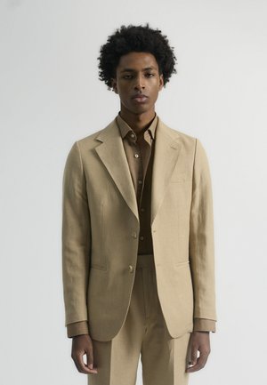 Traje de lino beige con blazer a medida y pantalones a juego, combinado con una camisa de botón marrón. Textura suave, corte moderno, detalles mínimos.